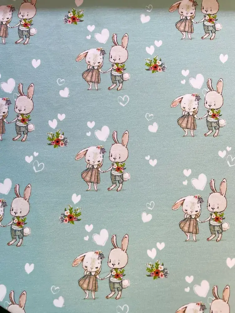 Weicher Jersey-Stoff für Kinder in Mintgrün, bedruckt mit einem fortlaufenden Muster aus süßen Hasenpärchen, die sich an den Händen halten, kleinen Blumensträußen und weißen Herzen. Perfekter Kinderstoff als Meterware für Kleidung.