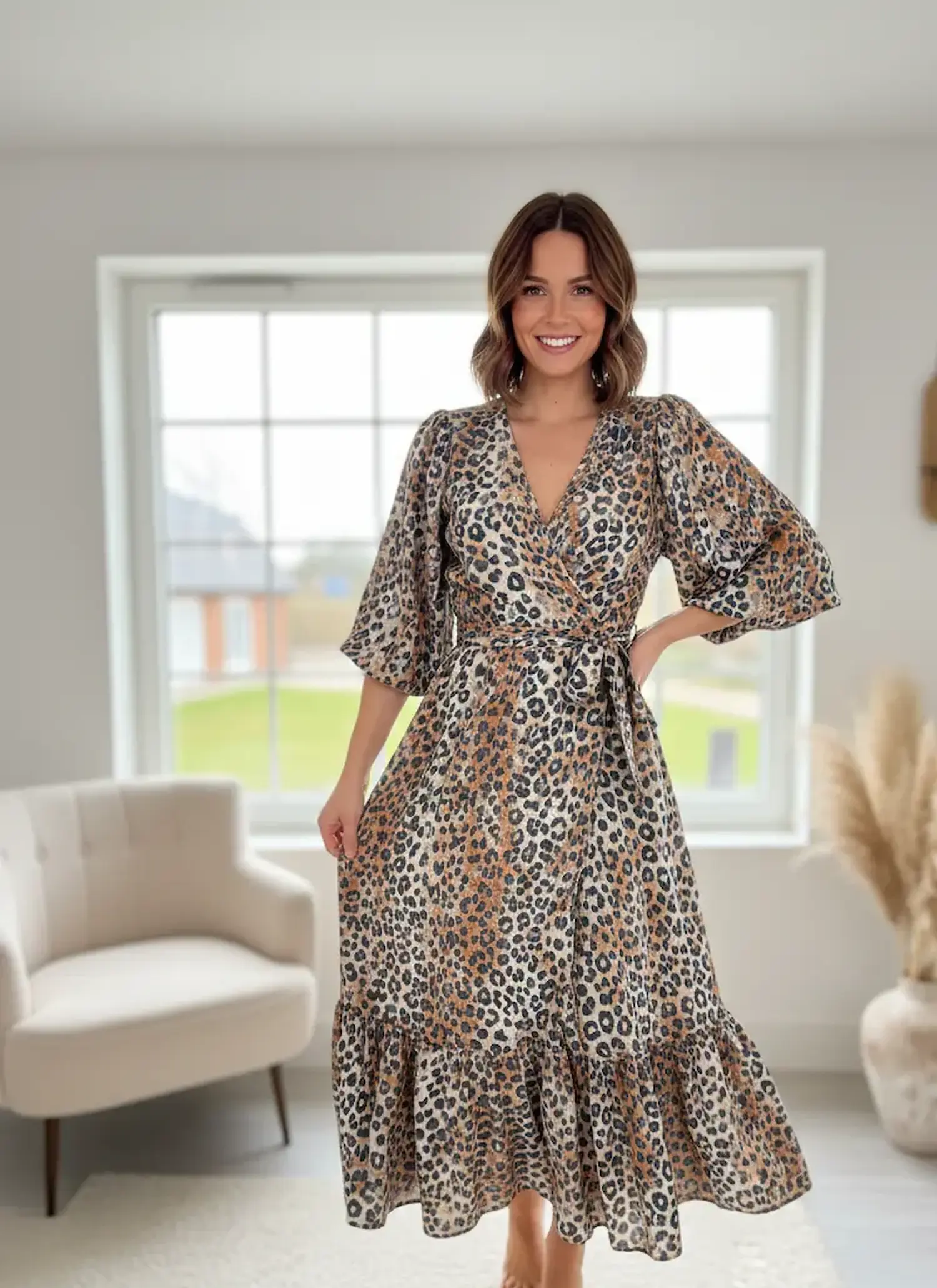 Eine lächelnde Frau präsentiert ein langes Wickelkleid aus luftigem Musselin-Stoff mit modernem Leopardenmuster in Braun- und Beigetönen vor einem hellen Fenster.