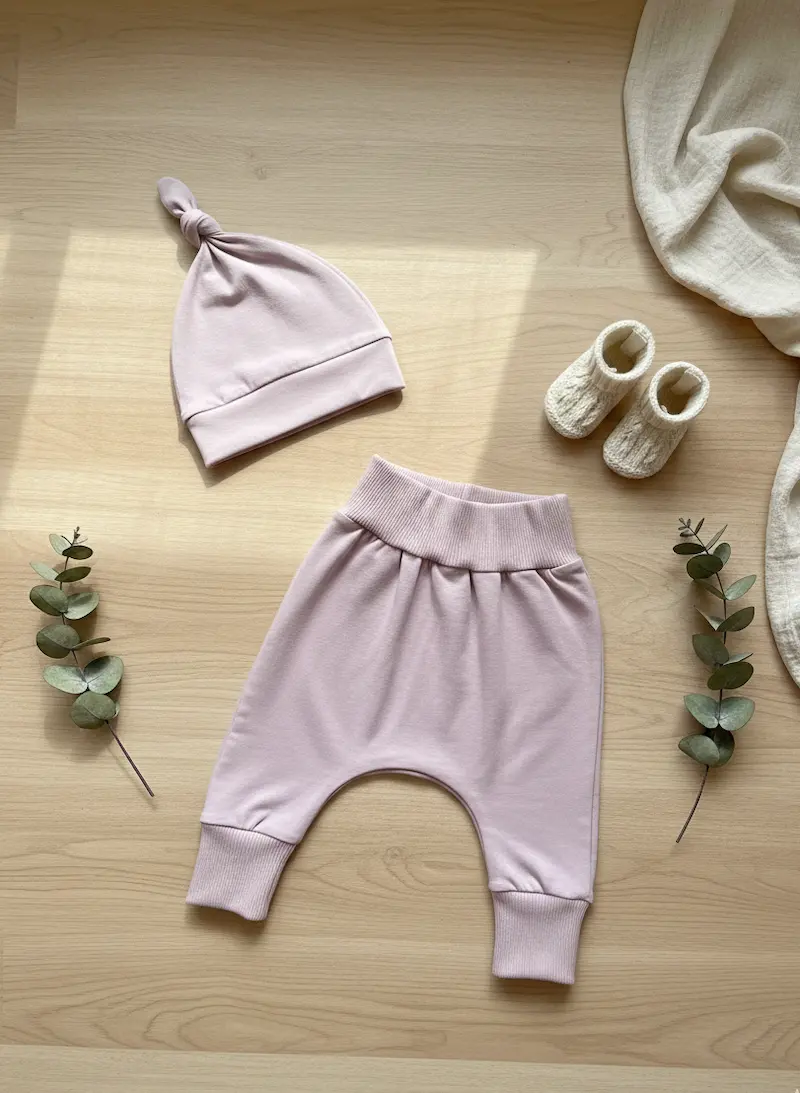 Baby-Set bestehend aus Knotenmütze und Pumphose aus fliederfarbenem Baumwolljersey, flach liegend auf hellem Holzgrund mit Eukalyptus-Deko.