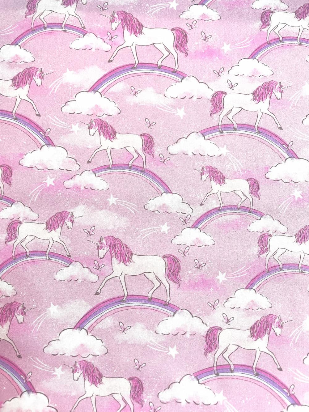 Draufsicht auf rosa Baumwoll-Webware als Meterware mit einem sich wiederholenden Motiv aus weißen Einhörnern, regenbogenfarbenen Bögen, weißen Wolken und kleinen Sternen.