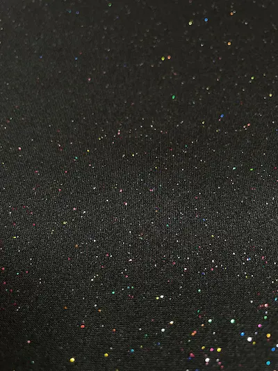 Nahaufnahme eines schwarzen Jerseystoffs, der mit vielen kleinen, bunten Glitzerpunkten (Multicolor) übersät ist und im Licht funkelt.