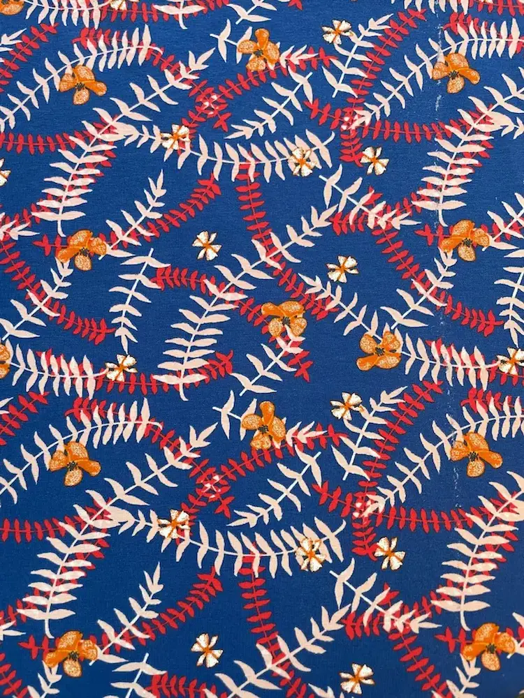 Blauer Baumwolljersey als Meterware bedruckt mit einem dichten, verschlungenen Muster aus hellbeige-rosa und roten Farnblättern sowie kleinen orangefarbenen und weißen Blüten.