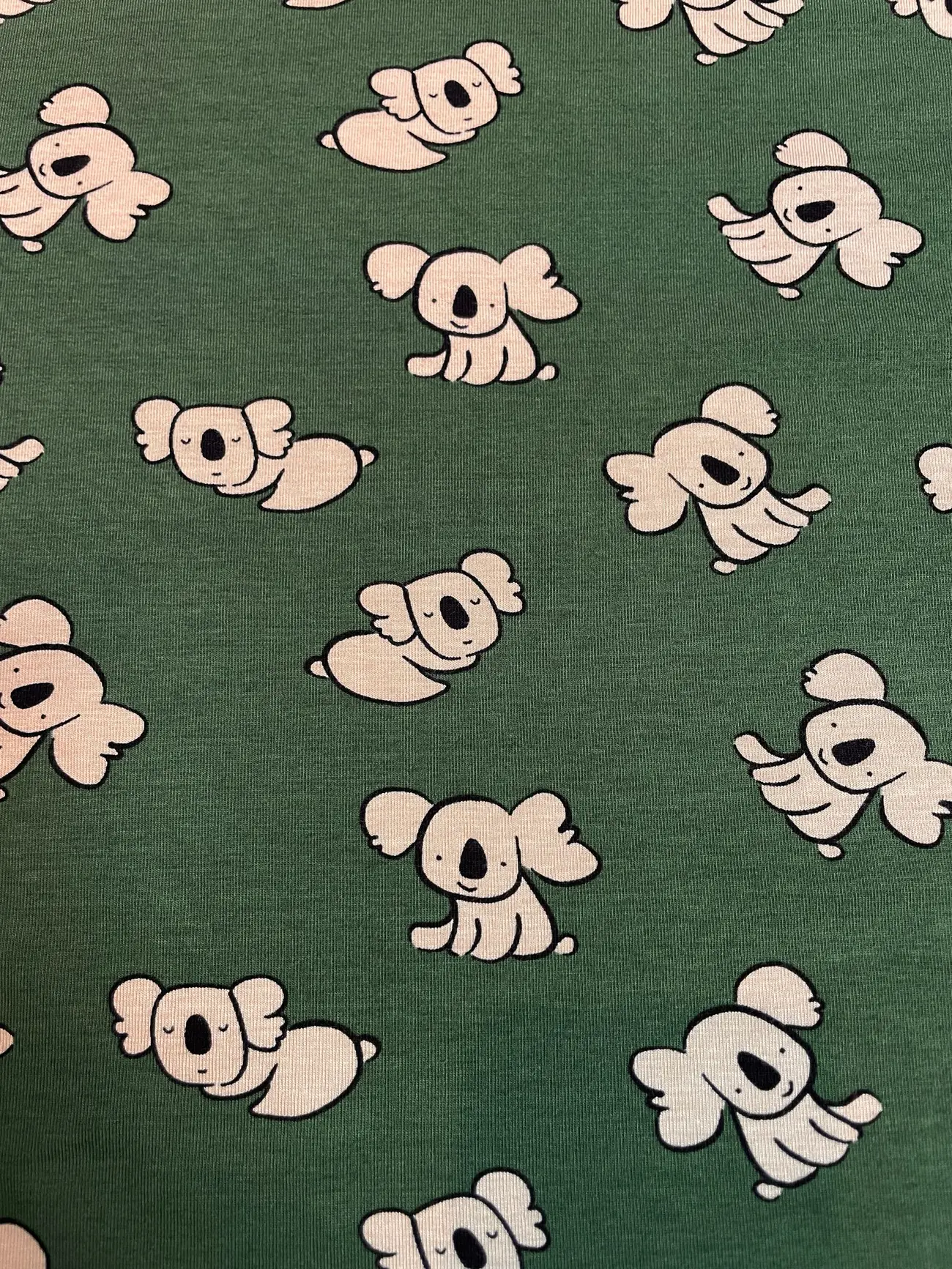 Ein weich drapierter Baumwolljersey-Stoff als Meterware in einem matten Dunkelgrün / Salbeigrün. Im Faltenwurf ist ein niedliches Kindermuster aus minimalistisch gezeichneten, hellen Koalas in verschiedenen sitzenden und schlafenden Positionen zu sehen.
