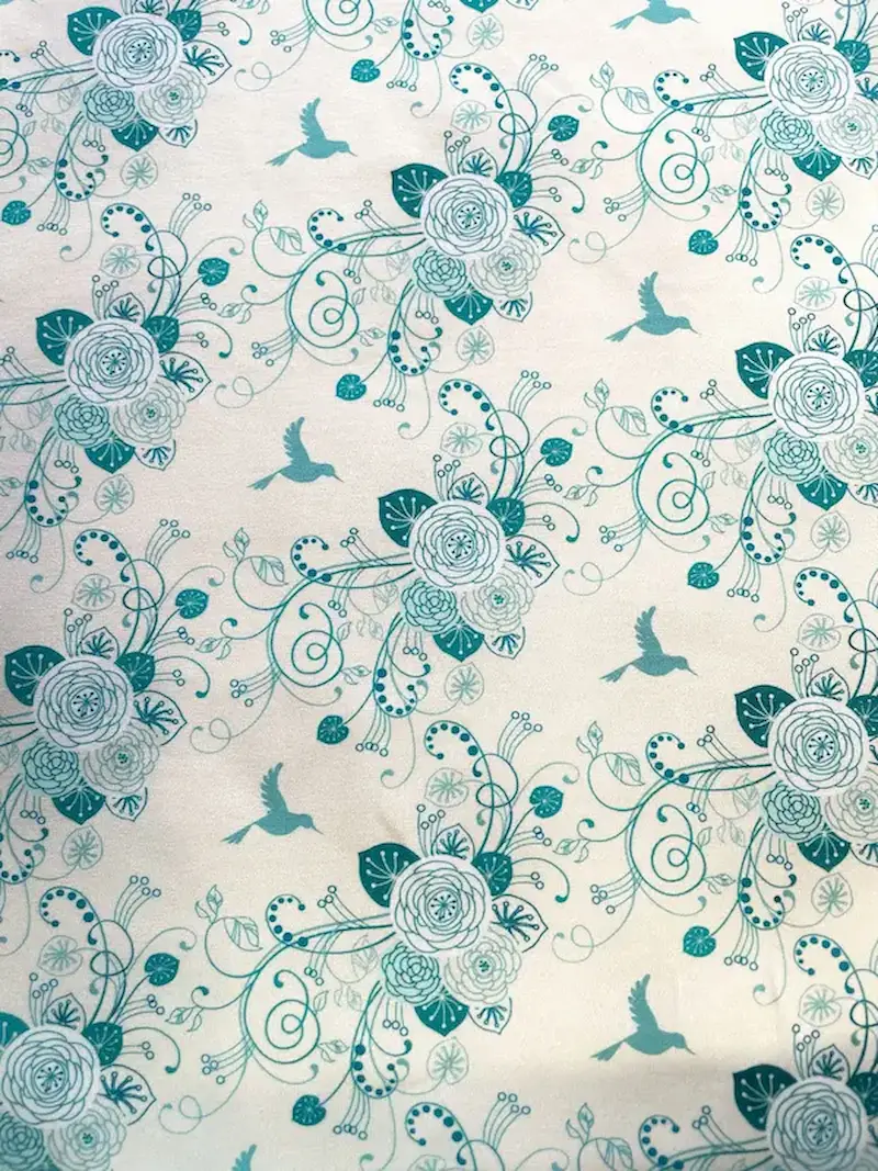 Hochwertiger, elastischer Baumwolljersey als Meterware mit weißem Hintergrund. Der Stoff ist mit einem filigranen, floralen Muster in verschiedenen Türkis- und Minttönen bedruckt, das fliegende Kolibris, kunstvolle Rosenblüten und verschnörkelte Blattrank