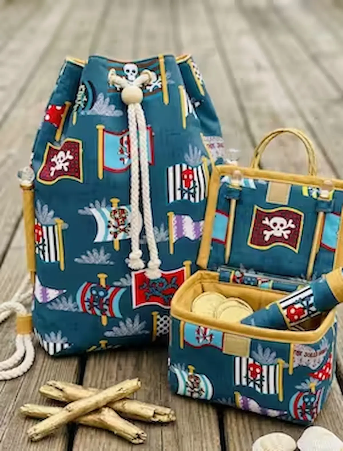 Genähtes Kinderzubehör aus petrolfarbenem Baumwoll-Webware-Stoff mit bunten Piratenflaggen, Totenköpfen und Wellenmotiven auf einem Holzsteg. Zu sehen sind ein großer Seesack-Rucksack mit dicker, weißer Kordel und eine kleine, aufklappbare Schatztruhe (Ut