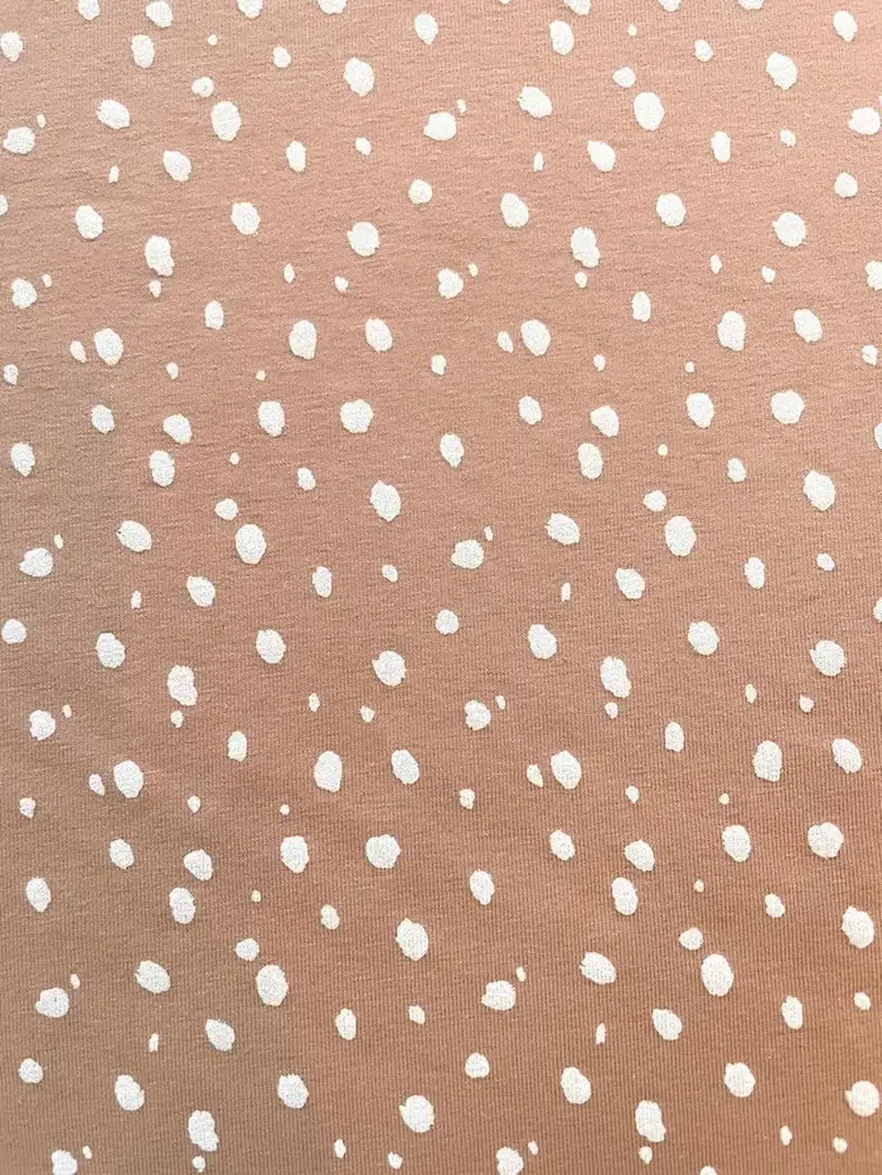 Draufsicht auf einen altrosa Jerseystoff mit unregelmäßig verteilten, handgezeichnet wirkenden weißen Punkten, die an ein dezentes Tiermuster erinnern.
