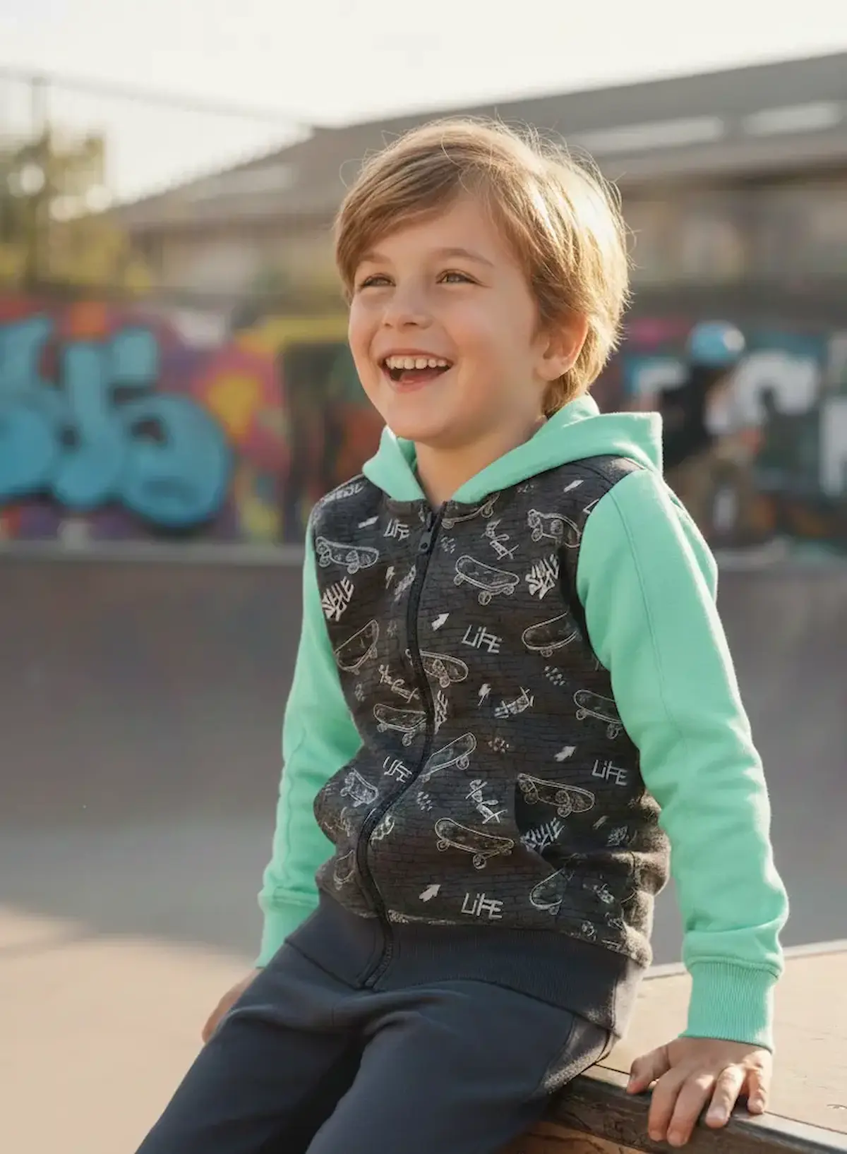 Ein lachendes Kind sitzt in einem Skatepark und trägt eine selbstgenähte Sweatjacke aus anthrazitfarbenem French Terry mit Skateboard-Motiven. Die Ärmel und die Kapuze sind farblich in hellem Mintgrün abgesetzt. Im Hintergrund ist eine Skate-Rampe mit Gra