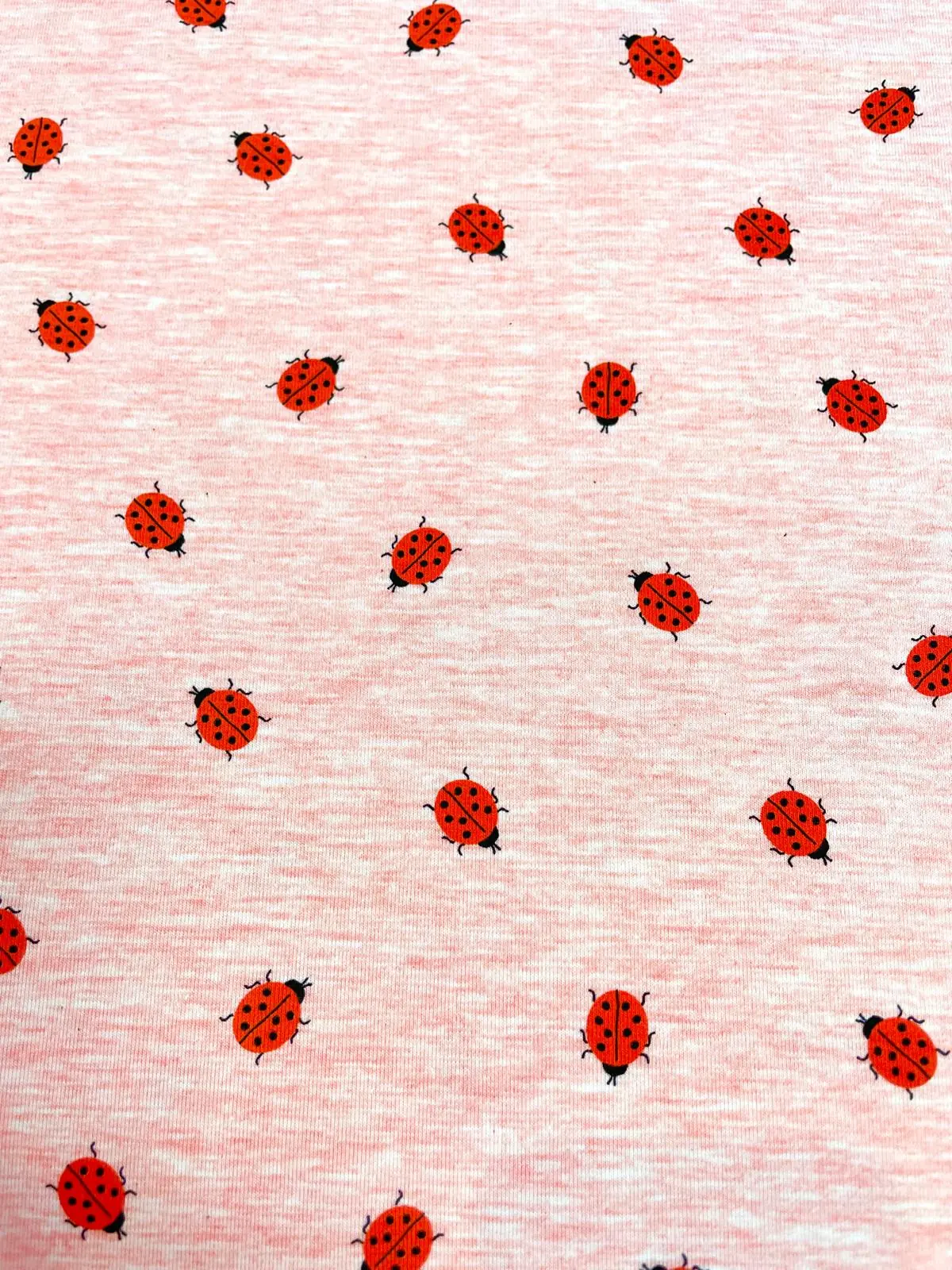 Süßer Baumwolljersey in hellem Rosa meliert, bedruckt mit kleinen roten Marienkäfern, ideal für Kinderkleidung, Baby-Accessoires und verspielte Nähprojekte.