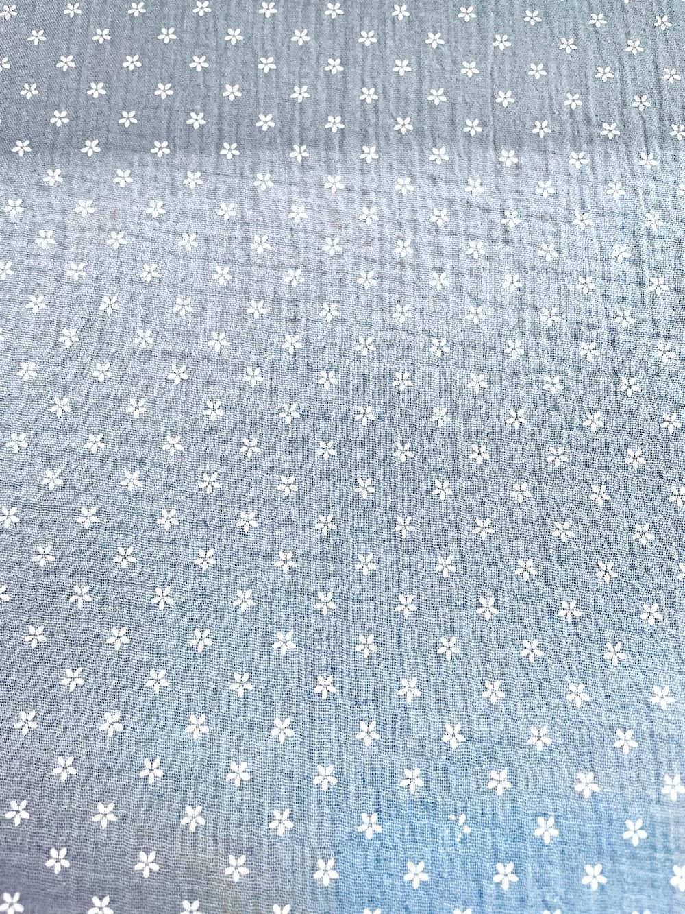 Flächenansicht von taubenblauem Musselin-Stoff mit einem Allover-Print aus kleinen weißen Blüten. Die Aufnahme zeigt die regelmäßige Musterverteilung und die charakteristische Crinkle-Struktur der Double-Gauze Meterware.