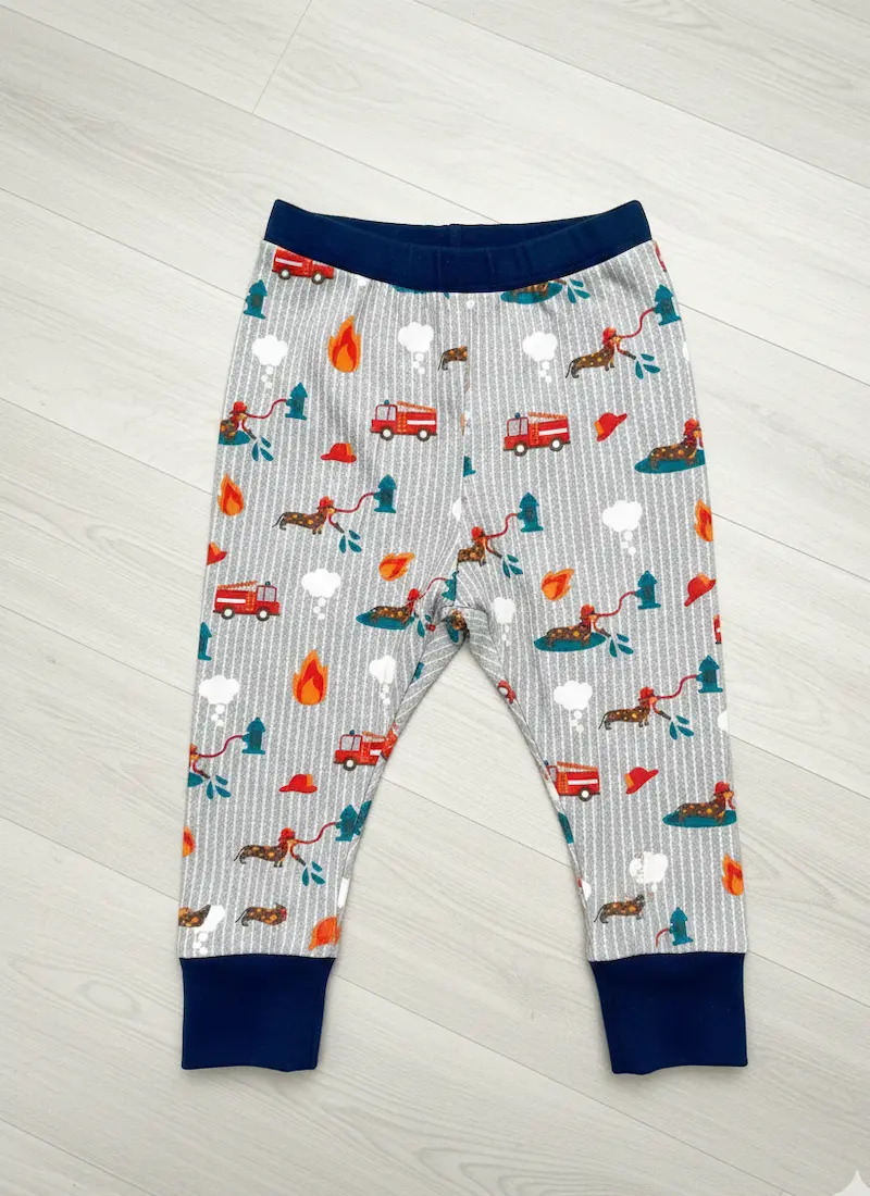 Eine selbstgenähte Kinderhose aus hellgrauem Baumwolljersey mit feinen weißen Streifen und einem bunten Feuerwehr-Motiv. Auf dem Stoff sind Dackel als Feuerwehrmänner, Einsatzwagen und Flammen zu sehen. Die Hose hat breite, dunkelblaue Bündchen an Bauch u
