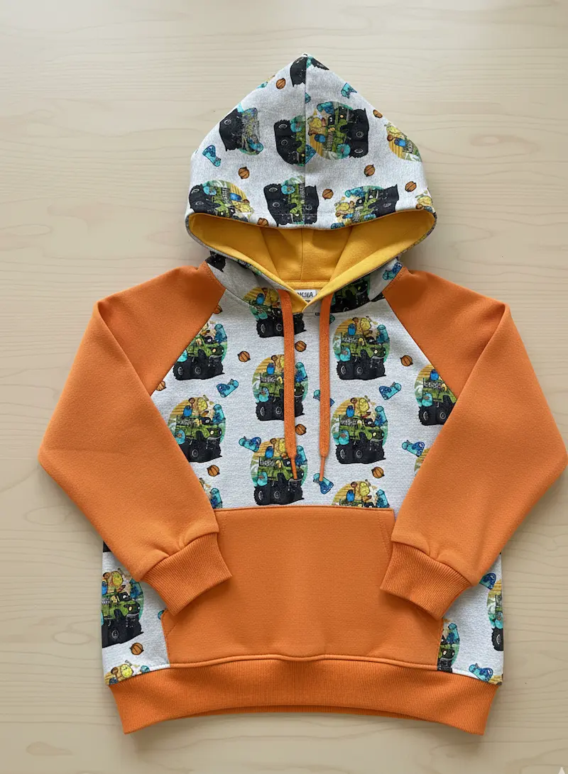 Selbstgenähter Raglan-Kinderhoodie aus grauem Monster-Truck-French-Terry mit Ärmeln und Tasche in Rostorange sowie gelbem Innenfutter.