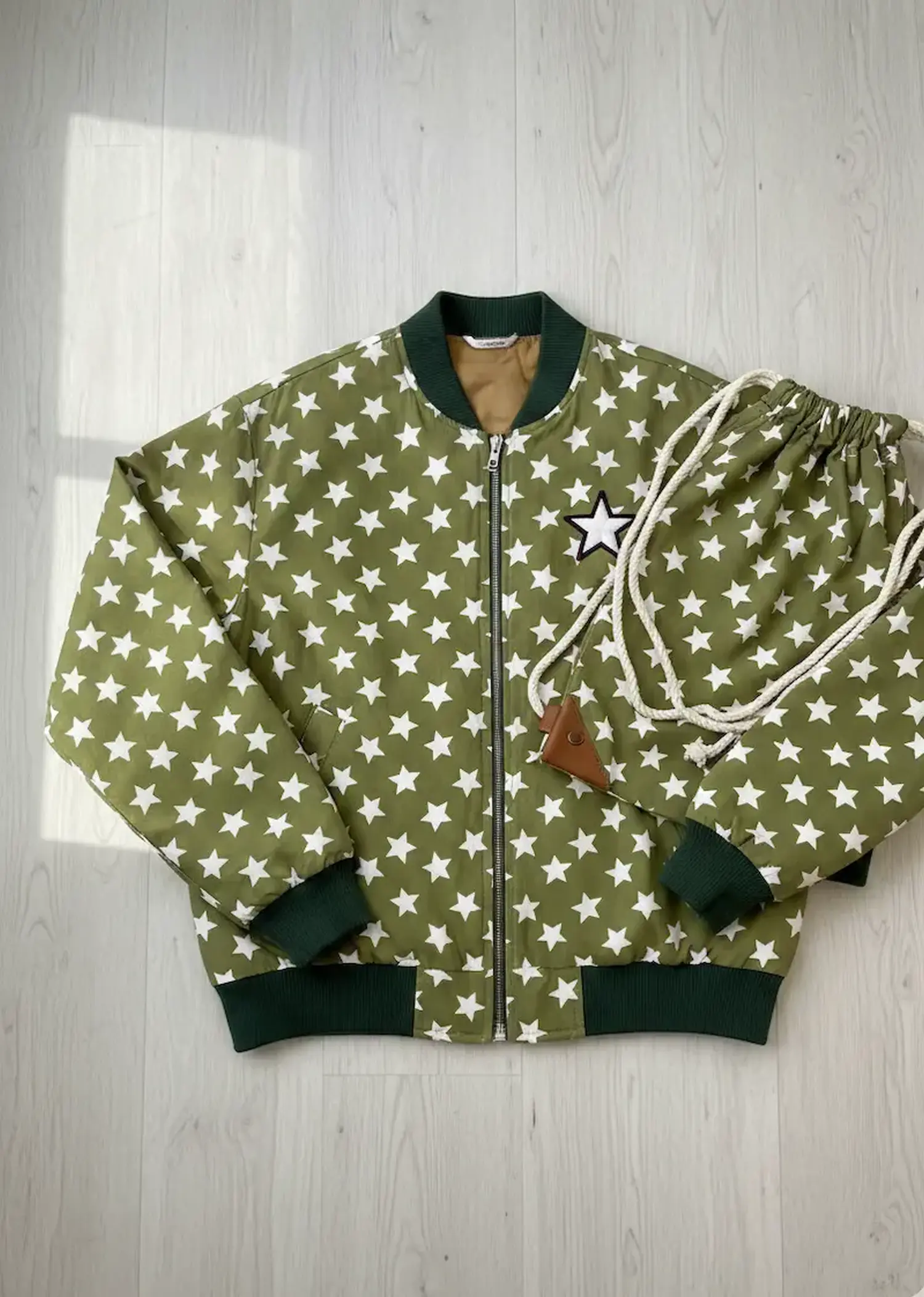 Nahaufnahme eines olivgrünen Jerseystoffs mit großen weißen Sternen sowie eine daraus genähte Bomberjacke mit passendem Turnbeutel auf hellem Holzboden.