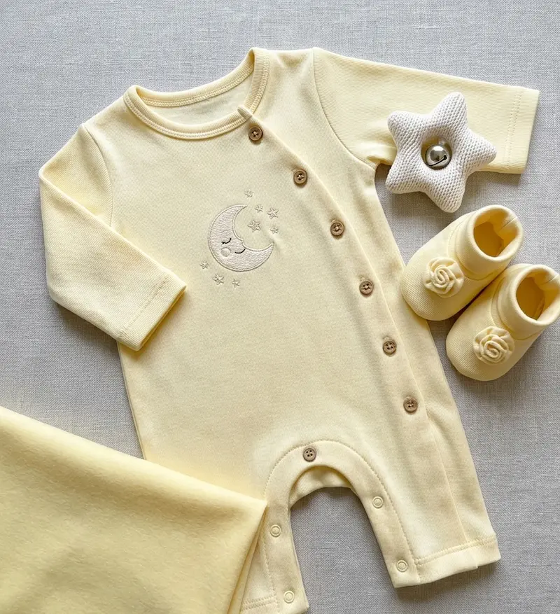 Hochwertiger Baumwolljersey in Pastellgelb als Meterware, flach liegend mit genähtem Baby-Strampler, Schühchen und Stern-Rassel auf grauem Hintergrund.