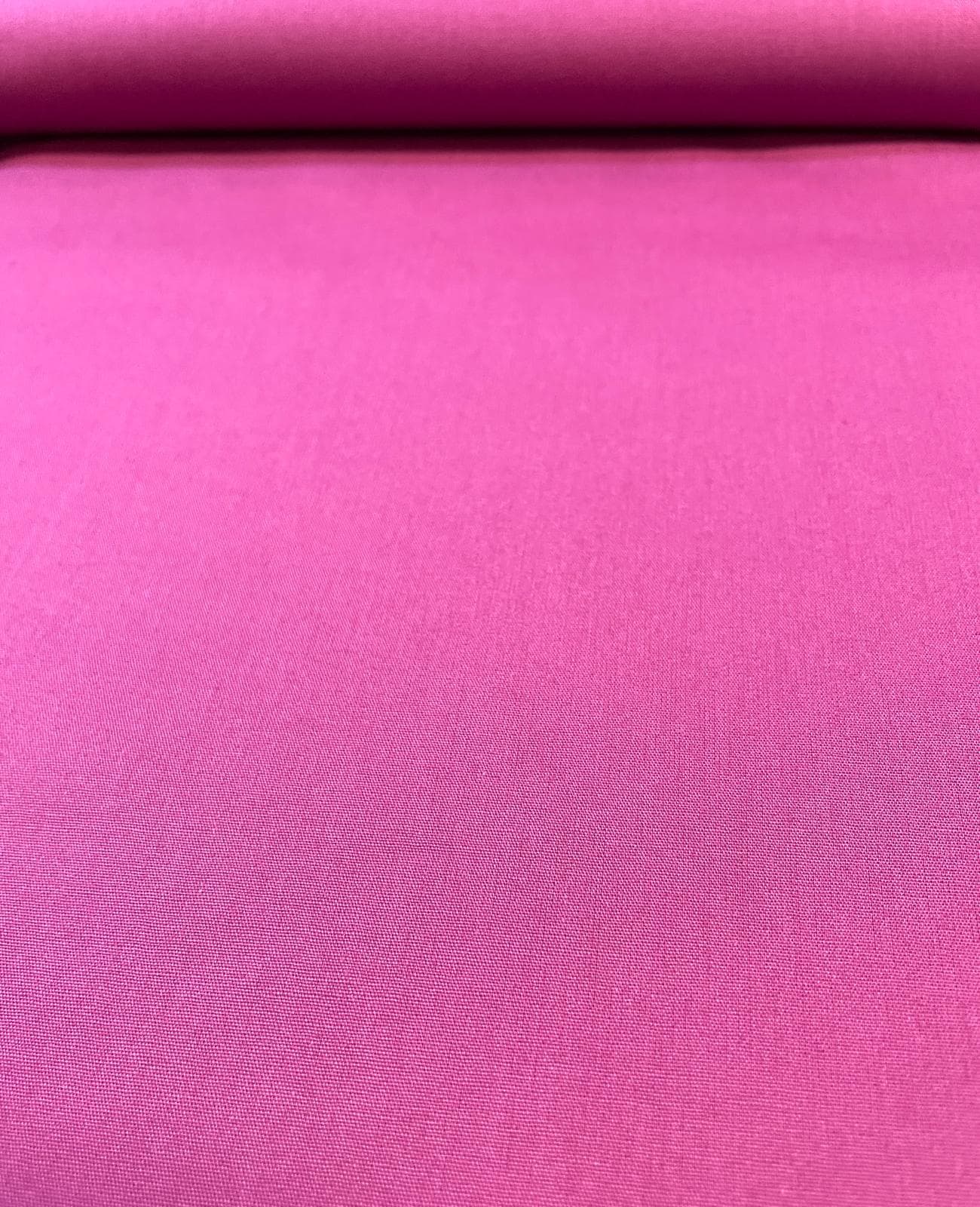 Gleichmäßig ausgebreiteter, unelastischer Baumwoll-Popeline Stoff als Meterware in kräftigem Magenta-Pink, der am oberen Rand glatt über eine Stoffrolle gewickelt ist und eine feine, dichte Webstruktur zeigt.