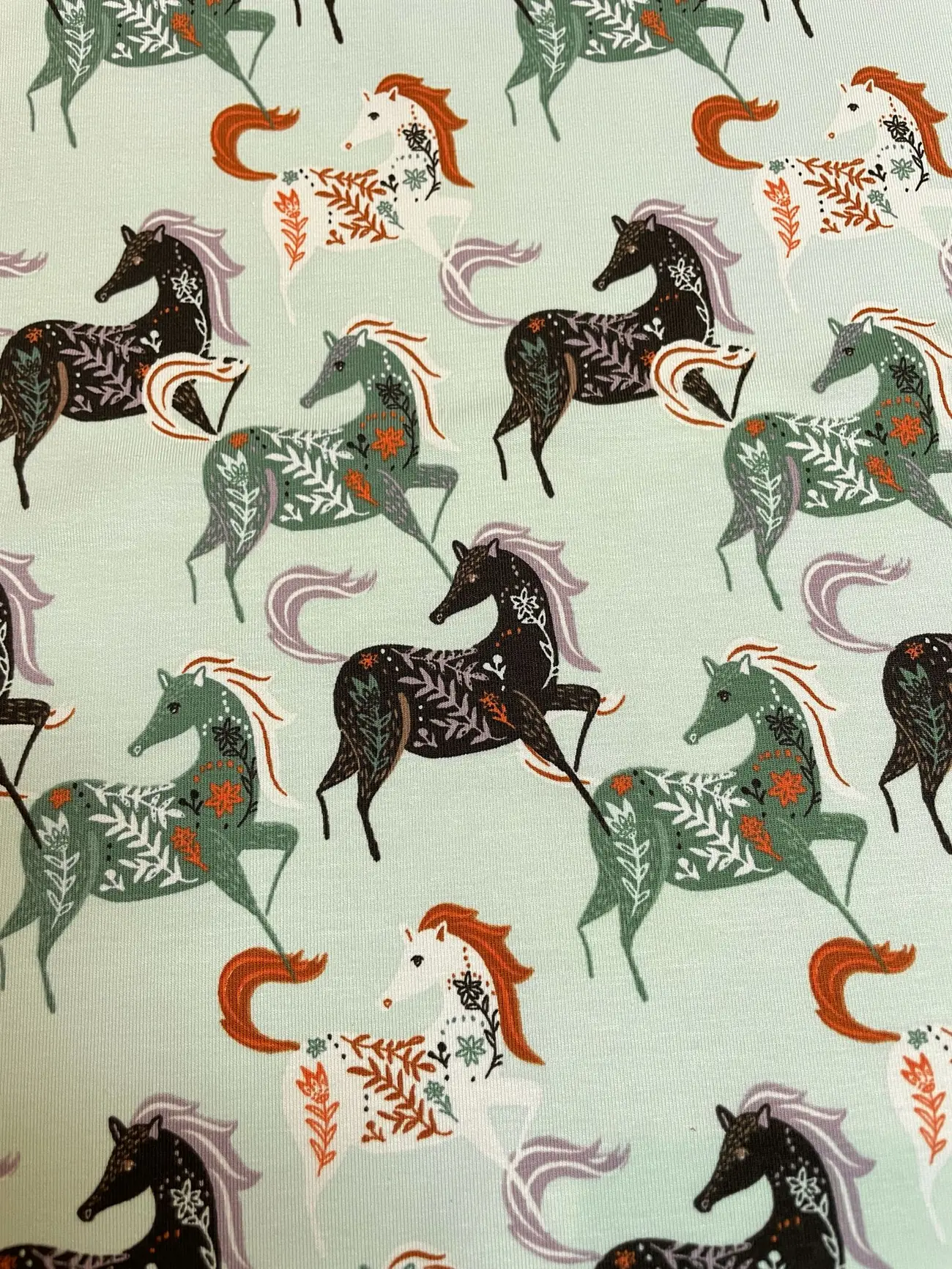 Weicher Jersey-Stoff in Mintgrün mit einem Muster aus galoppierenden Pferden in Weiß, Schwarz und Dunkelgrün. Die Pferde sind mit filigranen floralen Elementen, Farnen und herbstlichen Blättern im Folk-Art-Stil verziert.