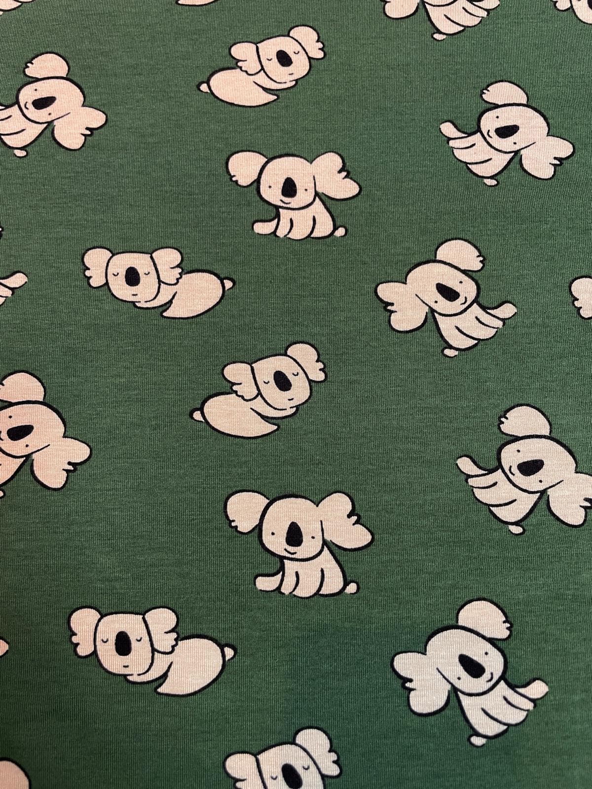 Ein flach liegender Bio-Baumwolljersey-Stoff als Meterware in einem matten Dunkelgrün / Salbeigrün, bedruckt mit einem schlichten Kindermuster aus kleinen, hellbeigen Koalabären, die teils sitzen und teils schlafen.