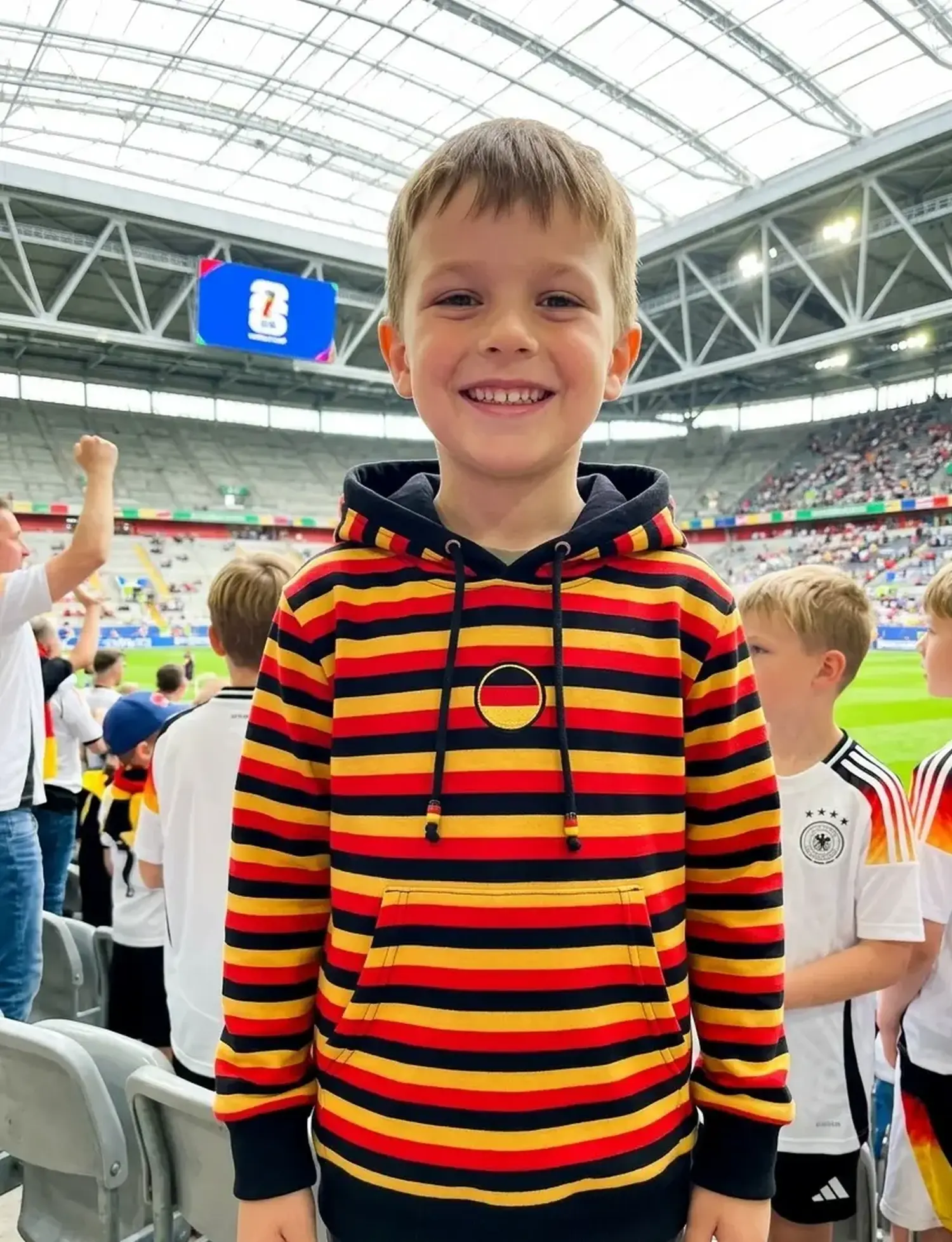 Ein lächelnder Junge im Stadion trägt einen selbstgenähten Kapuzenpullover aus gestreiftem Baumwolljersey in den Fanfarben Schwarz, Rot und Gold zur Fußball-WM.