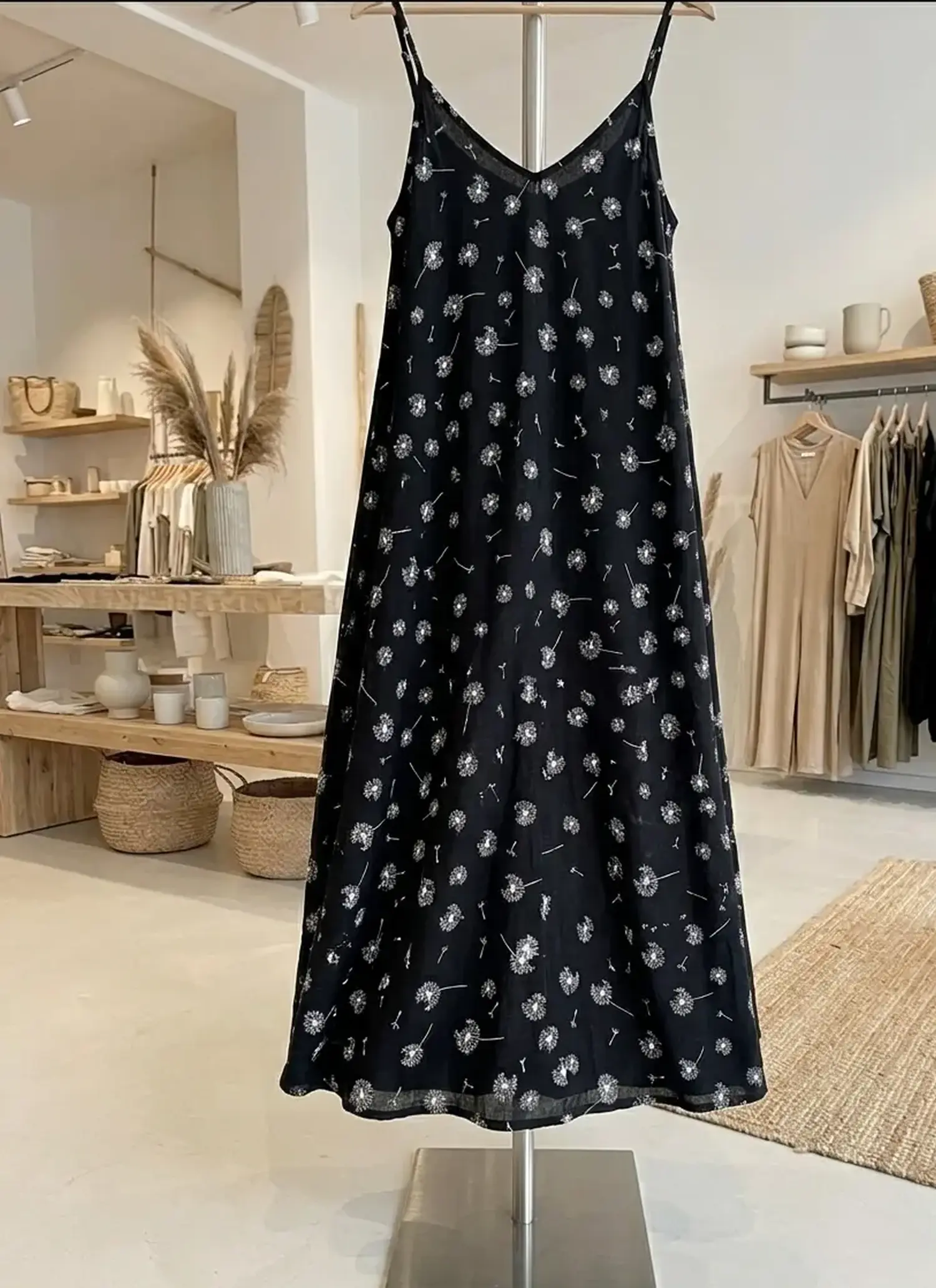Schwarzes Spaghettiträger-Kleid aus Viskose mit weißem Pusteblumen-Muster auf einem Kleiderständer in einer hellen Boutique.