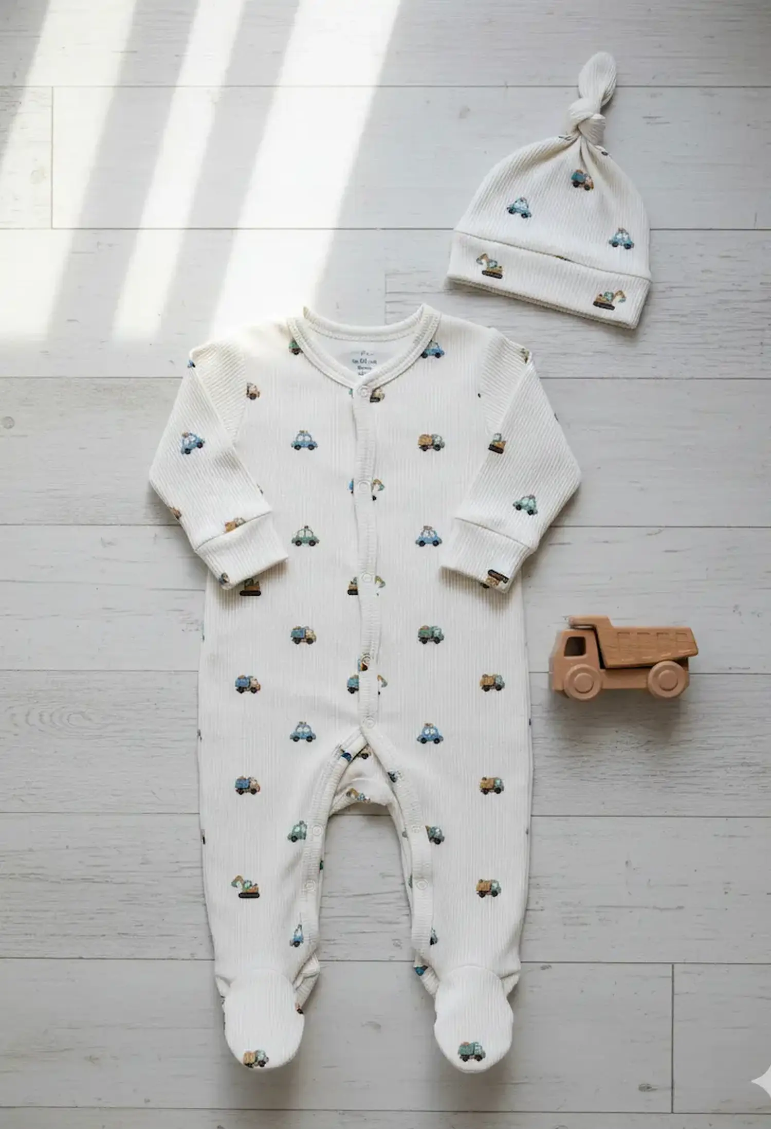 Ein genähter Baby-Schlafanzug mit Füßen und eine passende Knotenmütze aus weißem Rippjersey mit kleinen Autos und Lastern bedruckt, flach liegend auf hellem Holzboden neben einem Holz-Lkw.