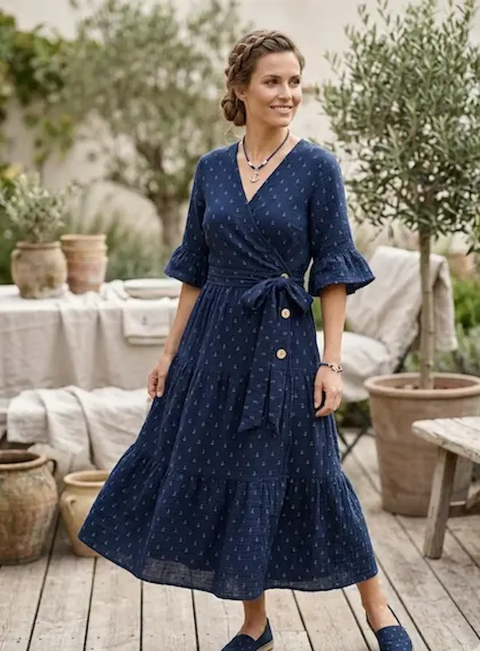Frau in einem gestuften, dunkelblauen Wickelkleid aus maritimem Musselinstoff mit kleinen Ankern, stehend auf einer Holzterrasse in einem Garten.