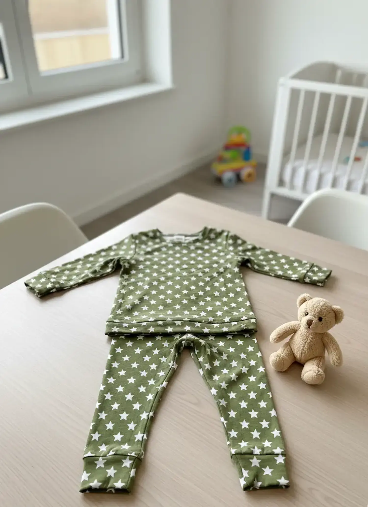 Ein handgenähter zweiteiliger Schlafanzug für Kinder aus olivgrünem Jerseystoff mit weißen Sternen, flach liegend auf einem Holztisch neben einem kleinen Teddybären im Kinderzimmer.