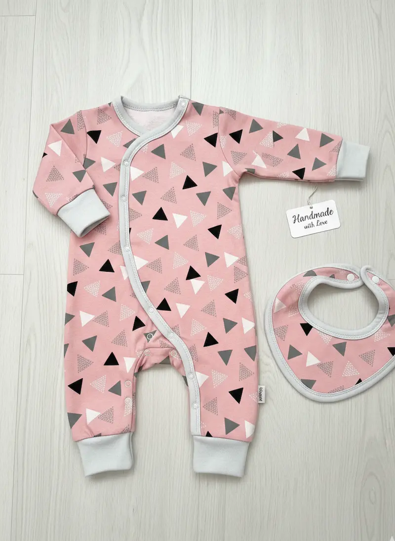 Genähtes Designbeispiel aus unserem rosafarbenen Jersey-Stoff mit Dreiecksmuster: Ein handgemachter Baby-Strampler (Einteiler) mit schräger Druckknopfleiste und hellgrauen Bündchen. Direkt daneben liegt ein passendes, kleines Halstuch bzw. Lätzchen und ei