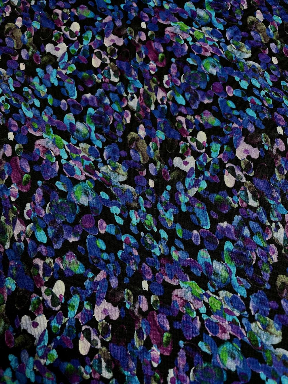 Kuscheliger Alpenfleece-Stoff als Meterware mit einem dunklen Untergrund und einem dichten, abstrakten Allover-Muster aus farbigen Tupfen, Flecken und Konfetti-Elementen in verschiedenen Nuancen von Blau, Lila, Violett und leuchtendem Grün.