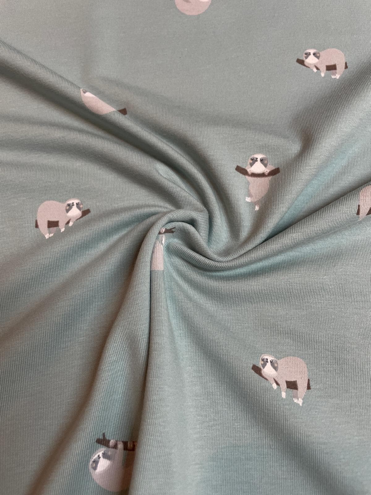 Mintgrüner Jersey-Stoff mit süßen Faultier-Motiven, die an Ästen hängen, weich drapiert zur Darstellung der Stoffqualität.