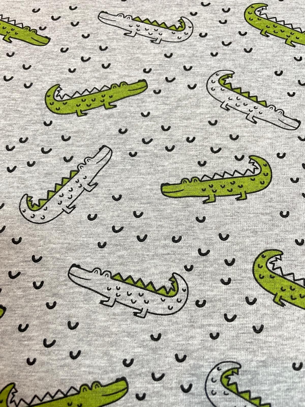 Kuscheliger Alpenfleece-Stoff in Grau-Melange mit Krokodil-Print. Niedliches Reptilien-Muster mit grünen Krokodilen und kleinen Wellen, ideal für warme Kinderkleidung und Decken.
