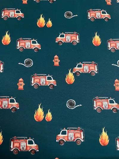 Wasserabweisender Softshell-Stoff mit Feuerwehrmotiven auf dunkelpetrolfarbenem Hintergrund. Das Muster zeigt rote Feuerwehrautos, lodernde Flammen, Hydranten und aufgerollte Feuerwehrschläuche. Ein detailreicher Outdoorstoff für mutige kleine Alltagsheld