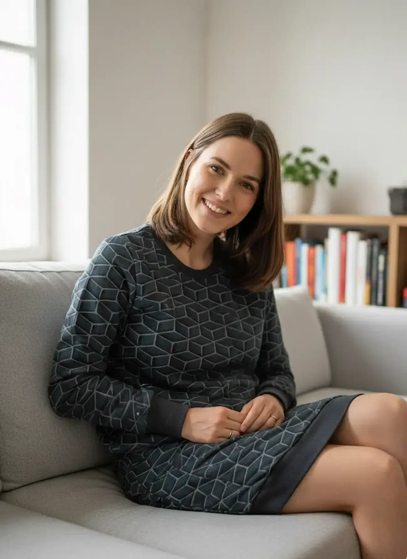 Eine lächelnde Frau sitzt auf einem grauen Sofa und trägt ein langärmeliges Kleid aus anthrazitfarbenem French Terry. Der Stoff hat ein modernes, geometrisches Wabenmuster in einem helleren Grauton. Das Kleid ist mit dunkelgrauen Bündchen an Ausschnitt un