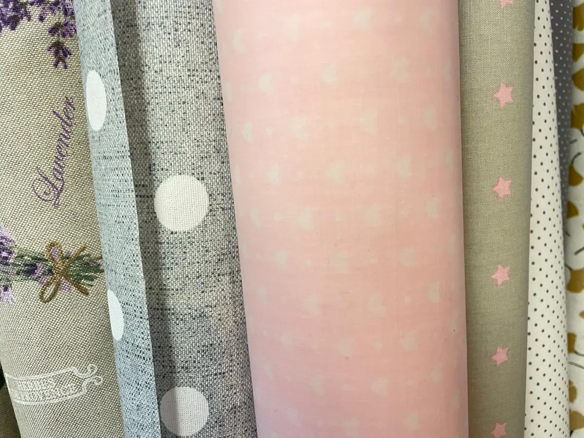 Sortiment an beschichteten Baumwollstoffen, darunter rosa Herzen, graue Punkte, Lavendel-Print und Sternen-Design, Meterware-Rollen.