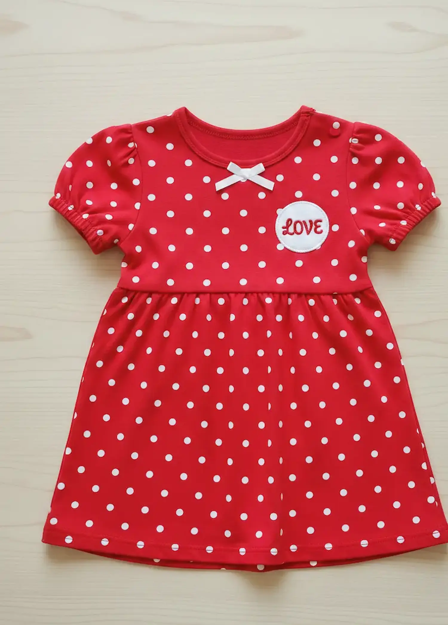Ein flach liegendes Babykleid aus rotem Jerseystoff mit gleichmäßigen weißen Punkten, Puffärmeln, einer kleinen weißen Schleife und einem runden Aufnäher mit der Aufschrift "LOVE".