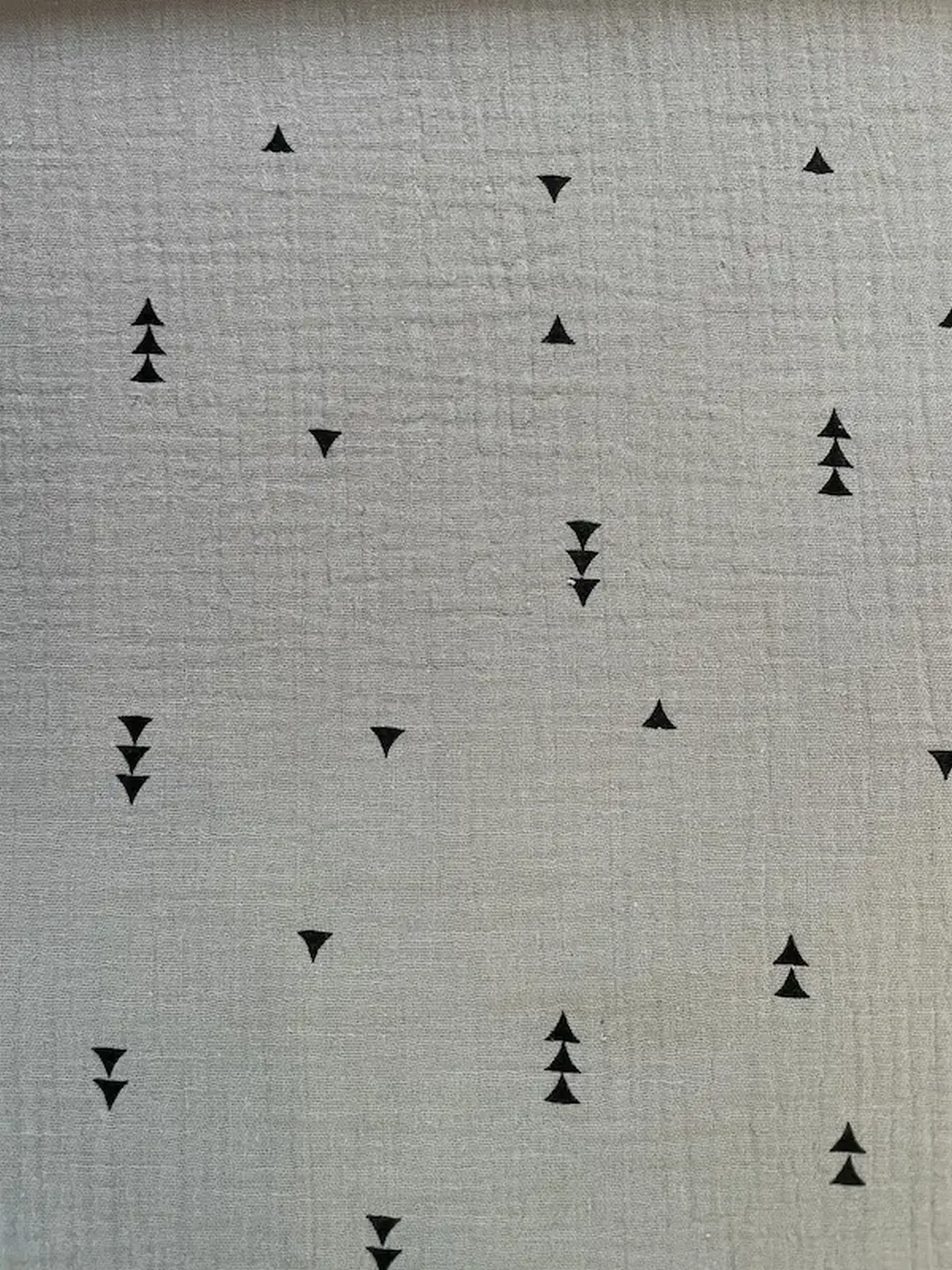 Leichter, luftiger Musselin-Stoff (Double Gauze) als Meterware in hellem Grau. Der Stoff ist mit einem dezenten, minimalistischen Muster aus kleinen, geometrischen schwarzen Dreiecken bedruckt. Die typisch gecrashte und weiche Gewebestruktur ist gut zu er