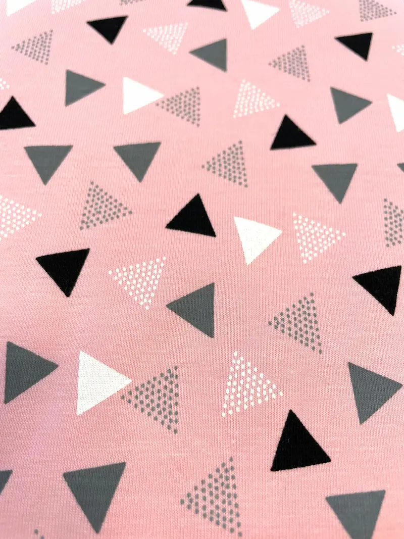 Hochwertiger Baumwolljersey als Meterware in zartem Rosa / Altrosa mit einem geometrischen Muster aus verschiedenen Dreiecken. Die Dreiecke sind abwechselnd in Schwarz, Dunkelgrau und Weiß gehalten, ergänzt durch Dreiecke aus feinen weißen und grauen Pünk