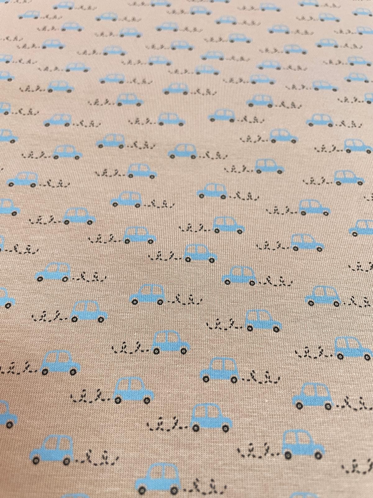 Beigefarbener Jerseystoff als Meterware mit einem minimalistischen Muster aus kleinen hellblauen Autos und schwarzen Reifenspuren.