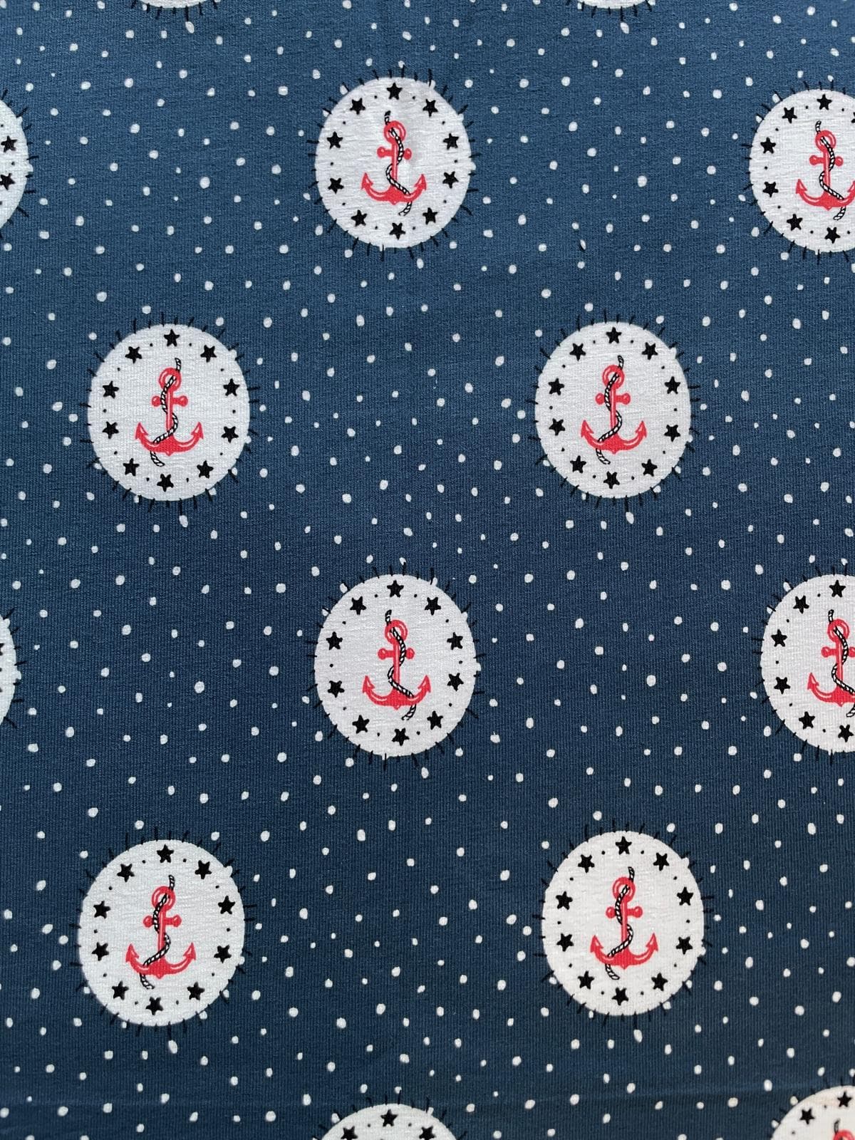 Nahaufnahme eines dunkelblauen Baumwolljersey-Stoffes mit kleinen weißen Punkten. Das Muster zeigt weiße Kreise, die mit schwarzen Sternen umrandet sind und in deren Mitte sich ein roter Anker mit Seil befindet.