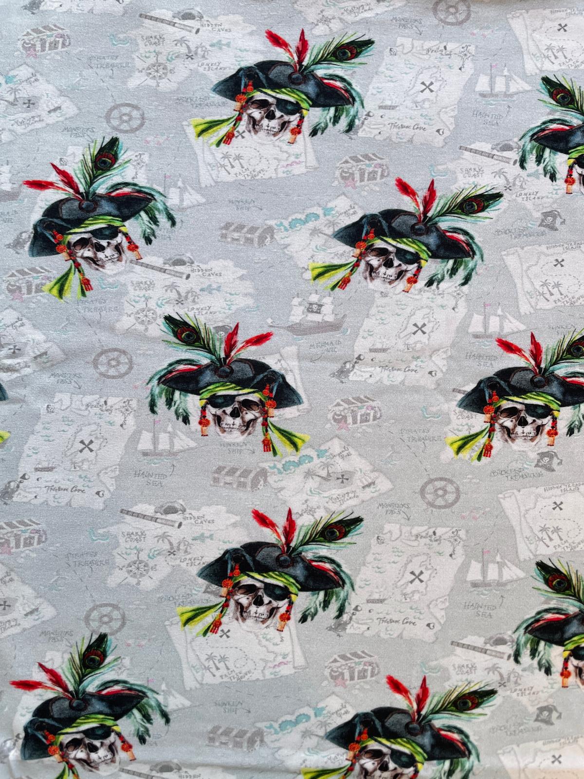 Hellgrauer Jersey-Stoff mit Piraten-Motiven, Totenköpfen mit roten Federn und einem Hintergrund im Vintage-Stil mit Landkarten-Fragmenten und Schriftzügen.