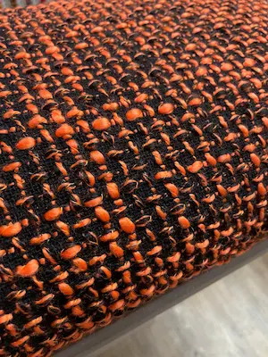 Ein hochwertiger Bouclé-Stoff mit einer ausgeprägten, groben Oberflächenstruktur. Das Gewebe zeigt ein lebendiges Melange-Muster aus leuchtendem Orange und tiefem Schwarz. Die charakteristischen Schlingen und Knoten des Materials verleihen dem Stoff eine