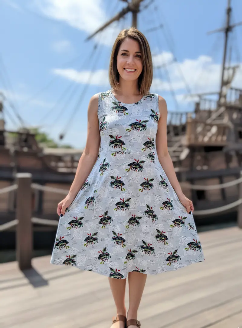 Eine Frau präsentiert ein knielanges Sommerkleid aus hellgrauem Jersey-Stoff mit maritimem Piraten-Print. Sie steht vor der Kulisse eines großen Holzschiffes. Der Stoff zeigt detaillierte Motive von Piratenschiffen und Schatzkarten.