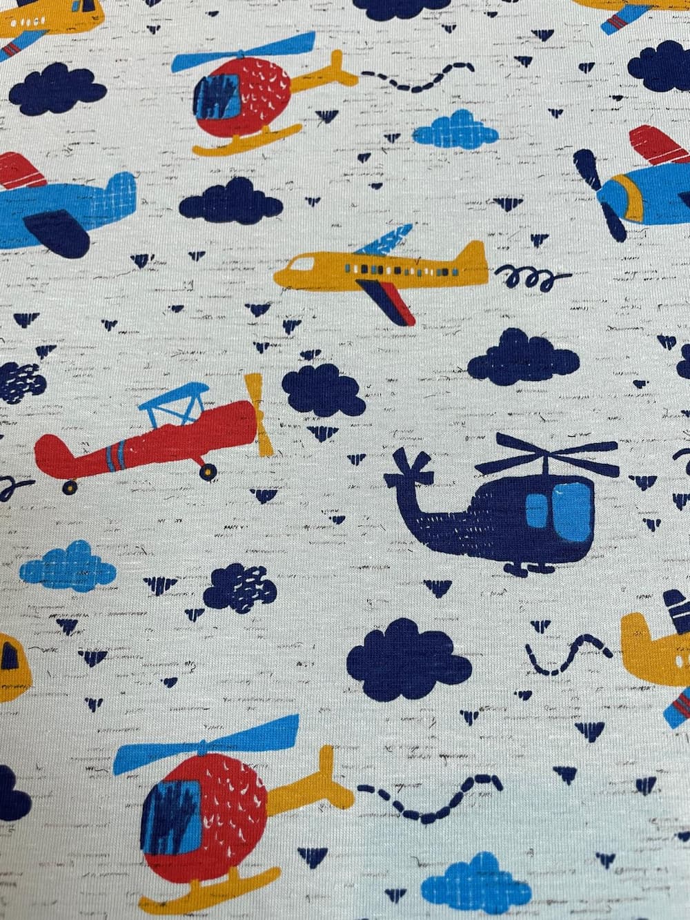 Ein flach liegender Baumwolljersey-Stoff als Meterware in Hellgrau mit Struktur-Optik, bedruckt mit einem farbenfrohen Kindermuster aus roten, blauen und gelben Flugzeugen, Hubschraubern und dunkelblauen Wolken.