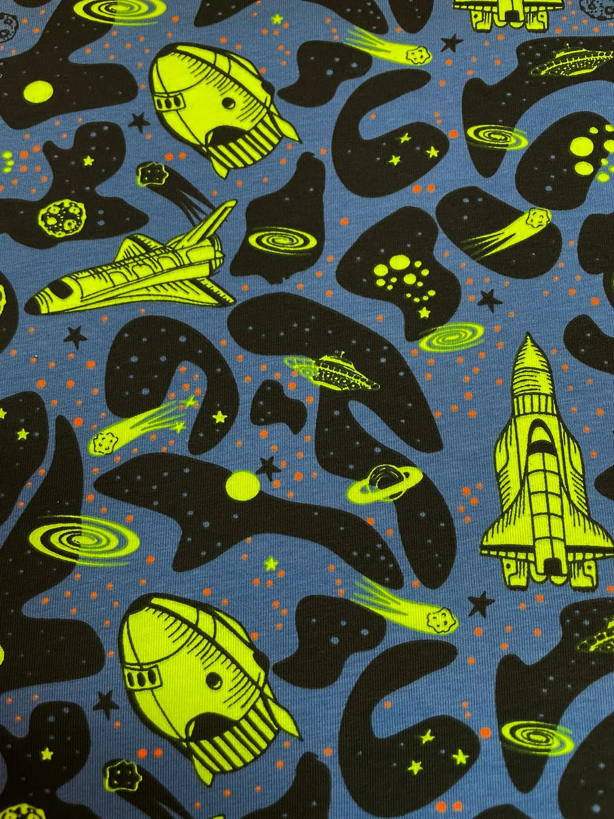 Ein flach liegender Baumwolljersey-Stoff als Meterware in Jeansblau, bedruckt mit einem coolen Weltall-Kindermuster aus neon-gelb-grünen Raketen, Space Shuttles, UFOs und Galaxien auf schwarzen Wolken-Formen mit kleinen orangefarbenen Sternenpunkten.