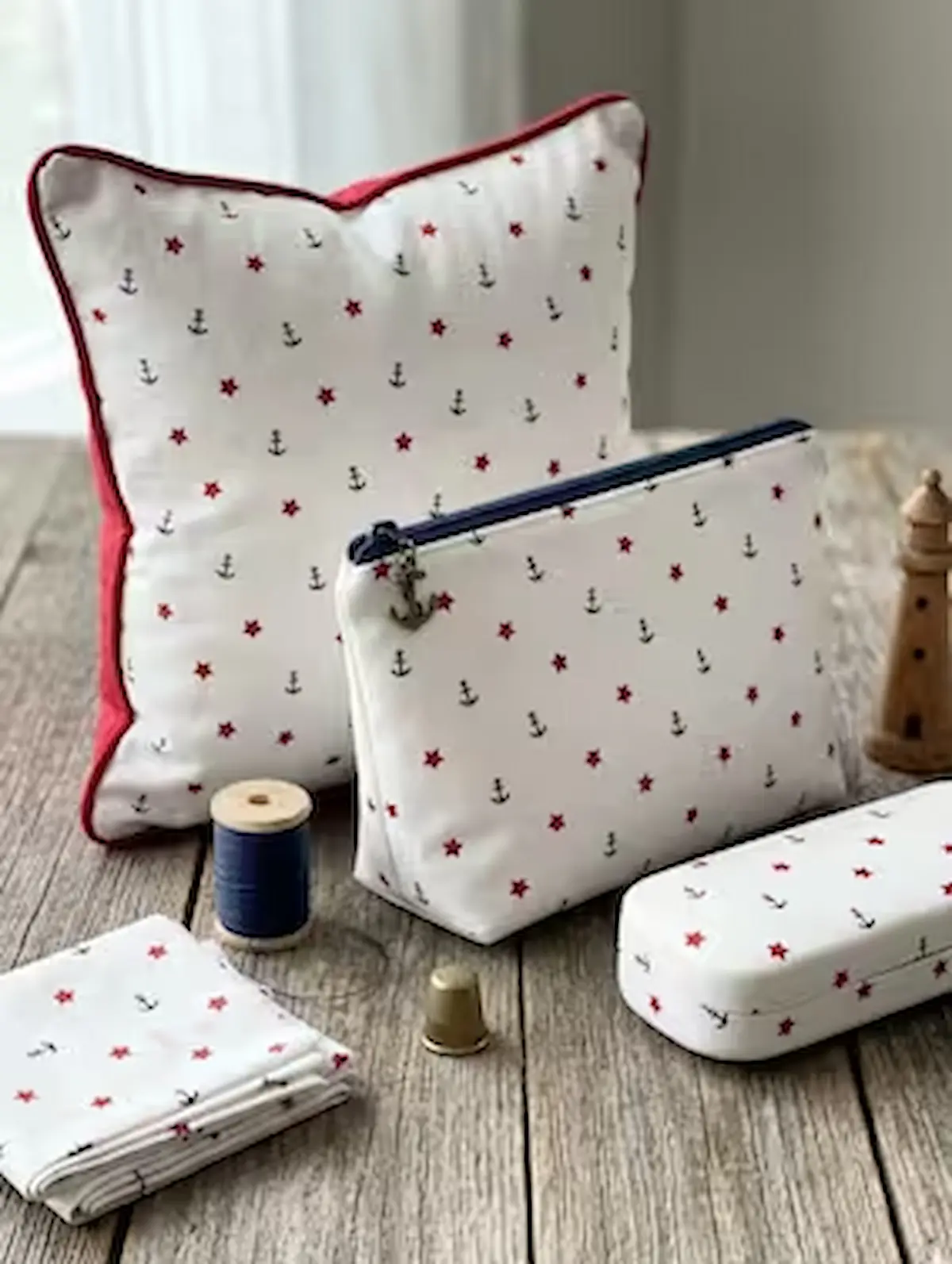 Genähte maritime Accessoires aus weißem Baumwoll-Webware-Stoff mit kleinen dunkelblauen Ankern und roten Sternen auf einem rustikalen Holztisch. Zu sehen sind ein Kissen mit roter Paspelierung, eine Kosmetiktasche mit Reißverschluss und Anker-Anhänger, ei