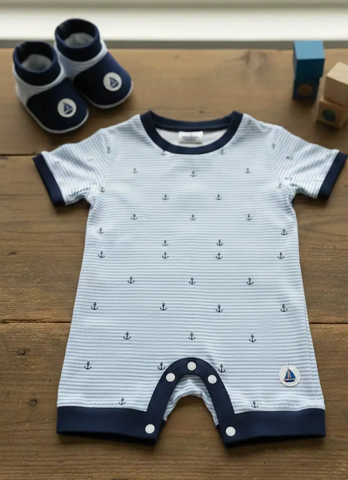 Ein handgenähter Baby-Strampler mit kurzen Ärmeln und Druckknöpfen im Schritt, gefertigt aus hellblau-weiß gestreiftem Baumwolljersey mit Anker-Motiv; dazu passende Babyschuhe auf einem Holztisch präsentiert als Inspiration für maritime Baby-Erstausstattu
