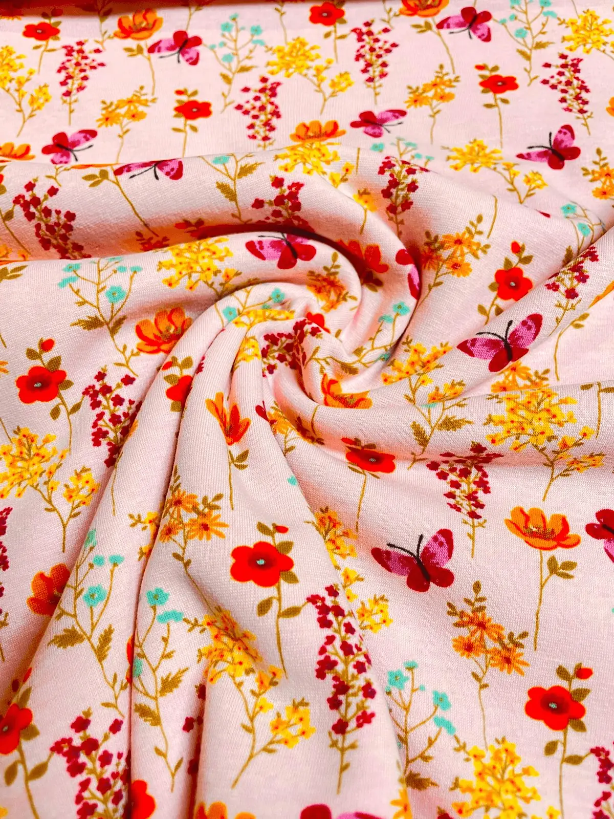 Kuscheliger Alpenfleece-Stoff als Meterware in Hellrosa mit einem Muster aus feinen Wildblumen in Orange, Gelb und Magenta sowie kleinen pinken Schmetterlingen.