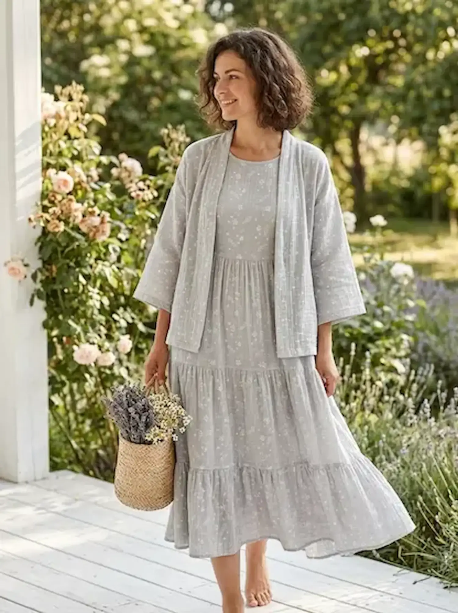Frau in einem hellgrauen Stufenkleid und passender Jacke aus Musselin-Stoff mit feinem weißen Blumenmuster im Garten.