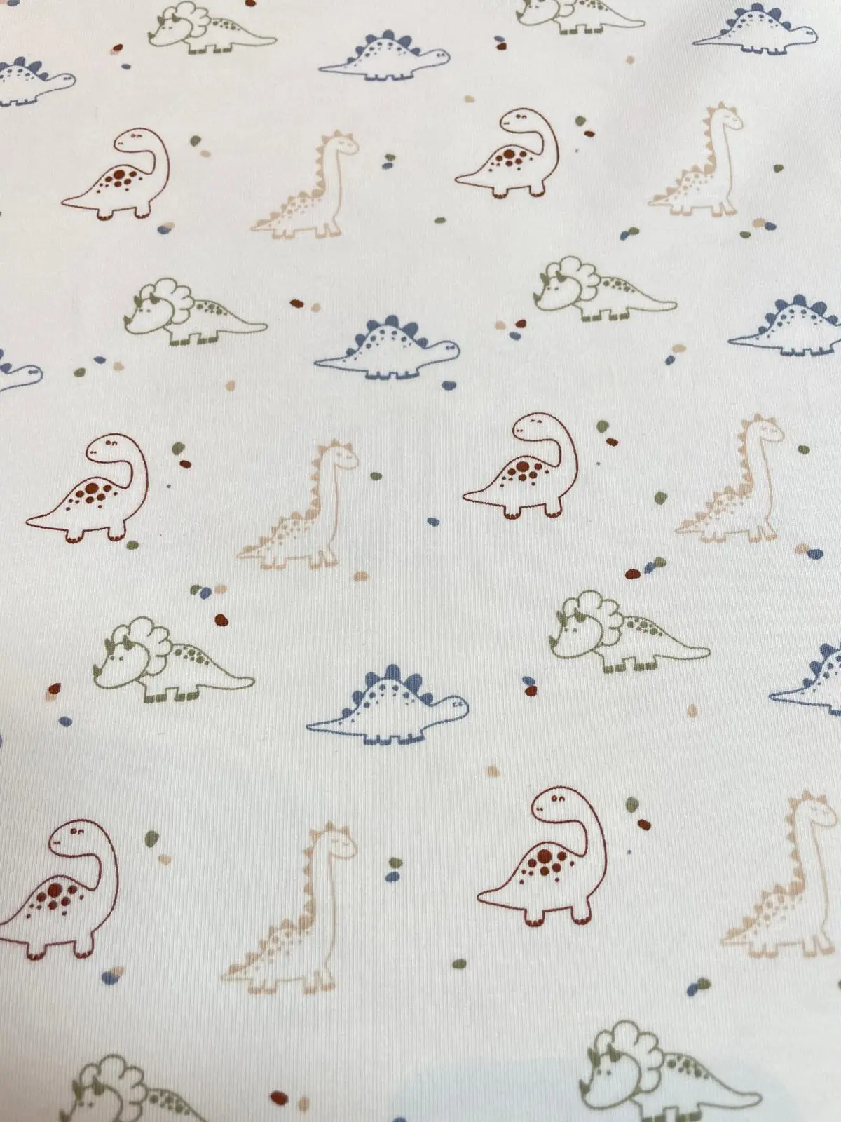 Weißer Jersey-Stoff mit kleinen Dinosaurier-Motiven wie Triceratops und Stegosaurus in Blau, Rostbraun und Beige mit bunten Sprenkeln.