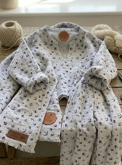 Helllila bis grau melierter Musselin-Stoff mit schwarzen Schwalben-Motiven als Meterware, verarbeitet zu einem hochwertigen Kinder-Set mit Leder-Details auf Holzuntergrund.