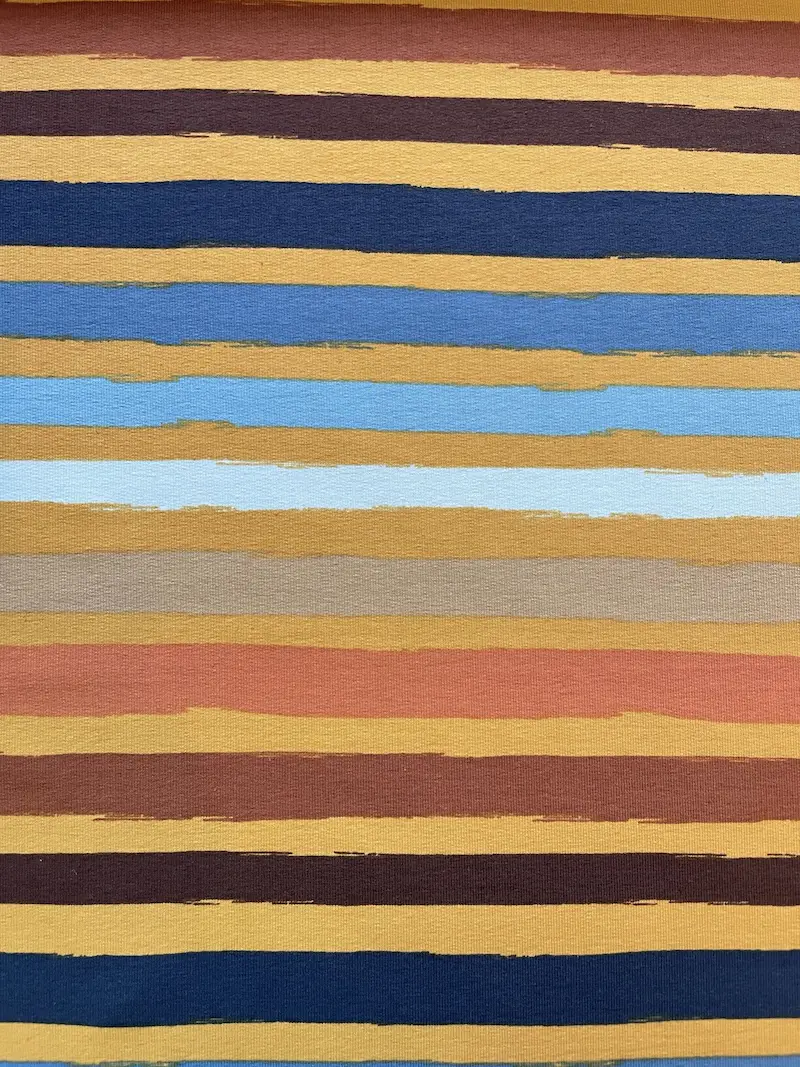 Hochauflösende Ansicht eines French Terry Sweatstoffs mit einem modernen, horizontalen Streifendesign. Die Streifen weisen eine künstlerische Pinselstrich-Optik auf und variieren in den Farben Senfgelb, Terracotta, Dunkelblau, Hellblau sowie Schokoladenbr