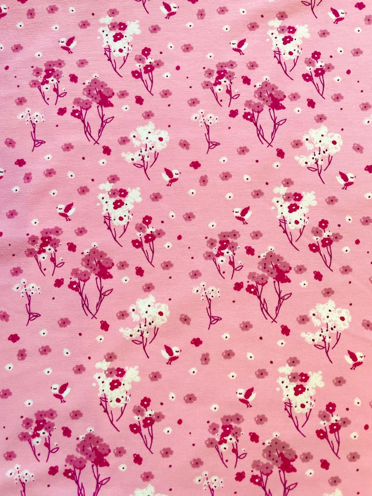 Detaillierte Nahaufnahme von hellrosa Baumwolljersey als Meterware mit einem verspielten Allover-Print aus kleinen, fliegenden Vögeln in Pink und Weiß sowie zarten Frühlingsblumen und Blütenzweigen.